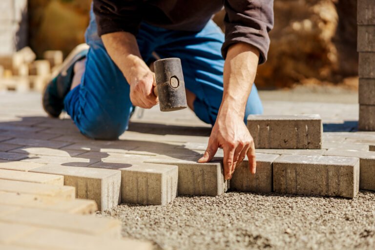 Young man laying gray concrete paving slabs in house courtyard on gravel foundation base. Master lays paving stones. Garden brick pathway paving by professional paver worker. Repairing sidewalk