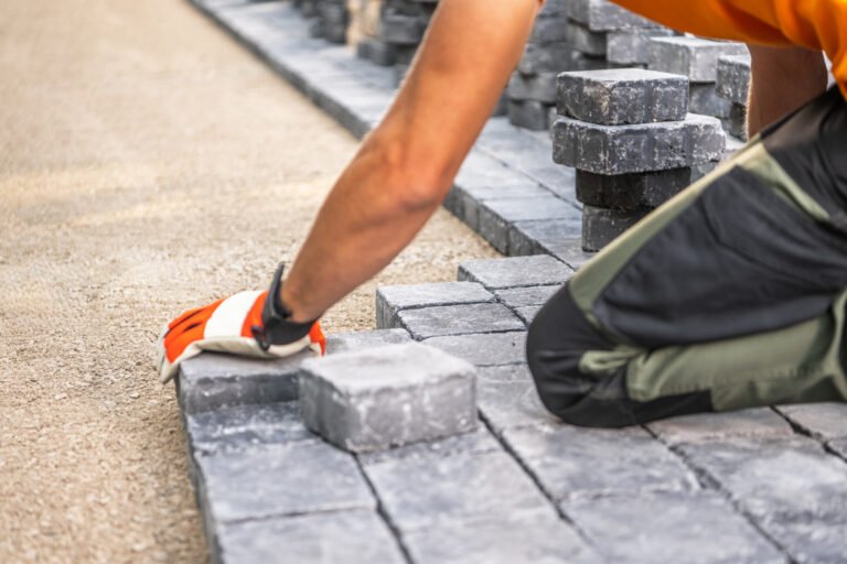 A worker carefully positions paving stones on a pathway, focusing on alignment and precision. Wearing gloves, the individual kneels on a gravel surface, surrounded by stacks of stone blocks. The bright daylight highlights the texture of the stones and the surrounding area.